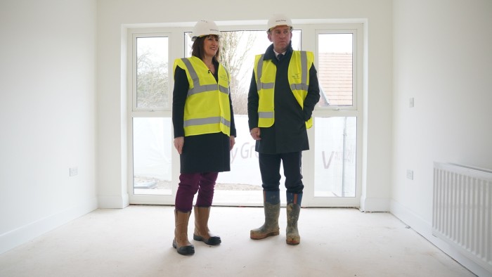 A man and a woman in hard hats and high-visibility vests survey a room in a newly built house