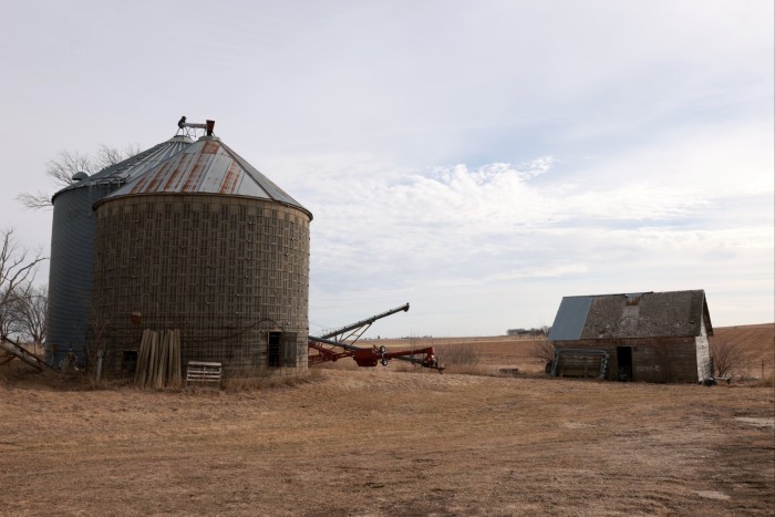 Dos silos de grano grande y un viejo cobertizo se sientan en un área seca y cubierta de hierba con amplios campos planos en el fondo bajo un cielo parcialmente nublado